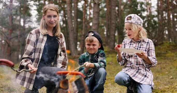mother with children frying and eating sausages by bonfire while camping in forest. family time, nature adventure