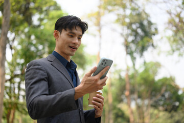 Handsome businessman with paper cup of coffee walking in city park and using smartphone