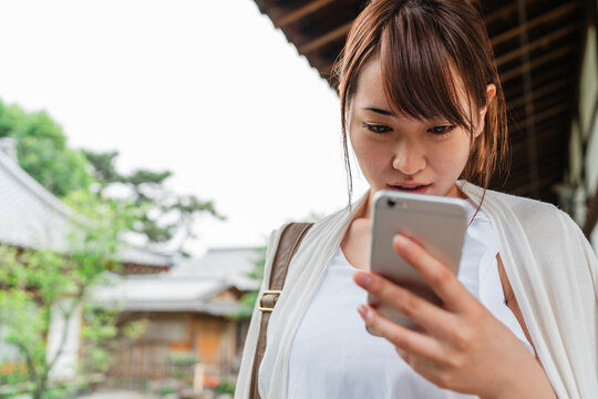 A young Japanese woman engages with her smartphone, her expression a blend of concentration and intrigue, against a backdrop of traditional Japanese architecture