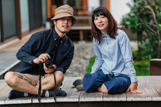 Two Japanese friends share a moment on a wooden deck; one with a camera in hand and the other with a warm smile, showcasing a casual, candid photography session in an outdoor setting