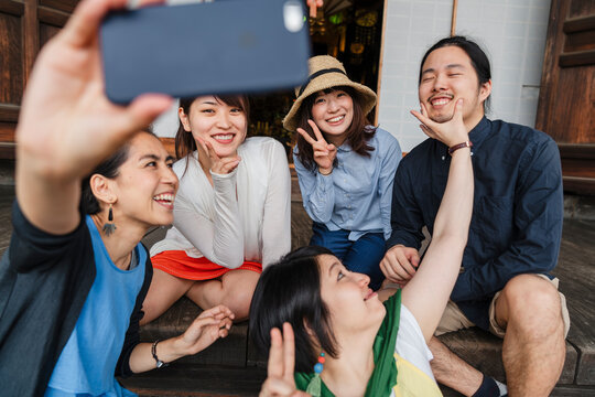 A group of five Japanese friends sharing a joyful moment while taking a selfie, sitting casually on wooden steps with one holding the phone