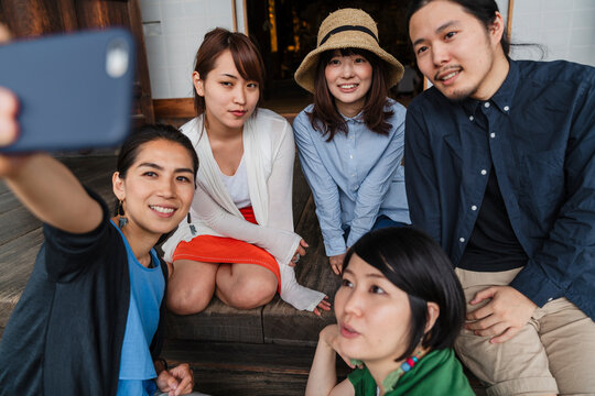 A group of five Japanese friends sharing a joyful moment while taking a selfie, sitting casually on wooden steps with one holding the phone