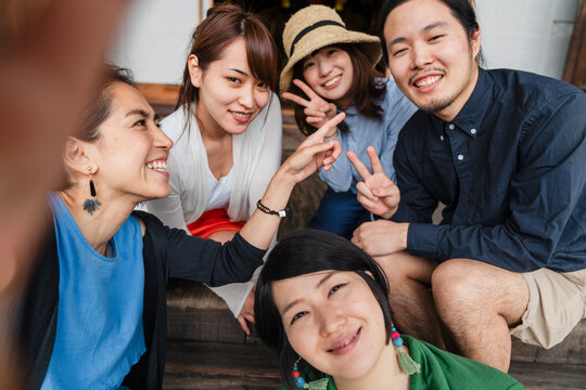 A group of five Japanese friends sharing a joyful moment while taking a selfie, sitting casually on wooden steps with one holding the phone