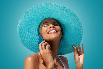 Happy woman portrait wearing blue sun hat and using mobile phone, isolated on turquoise bright background. Concept of summer beach holiday, shopping online, booking travel and resort accommodations