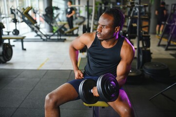 Fototapeta na wymiar Young African American man sitting and lifting a dumbbell close to the rack at gym
