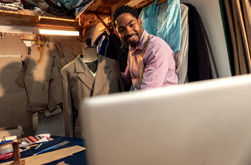 Male fashion tailor having video call with customer via laptop for the suit design in his workshop.