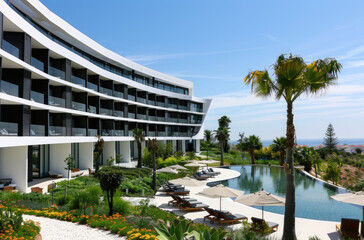 A large hotel with white walls and black windows, with curved architecture, a large pool in front of the building, a garden, palm trees - obrazy, fototapety, plakaty