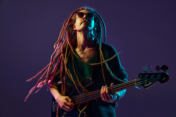 Captivating sight of man with dreadlocks, musician expressing his emotions through music, guitar playing against dark purple background in neon light. Concept of music, performance, festival, concert