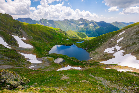 alpine landscape of fagaras mountains. capra lake of romania in summer. sunny weather with clouds on the blue sky. spots of snow and grass on the rocky hillside. stunning travel destination