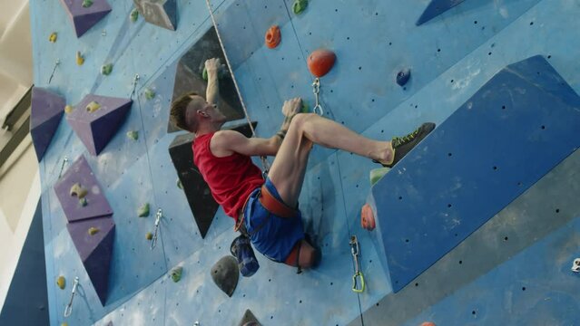 View from below of strong athlete with lower limb disability using rope while climbing wall in training gym