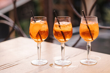 Three glasses with orange alcoholic drink on wooden table. Black straw in a drink. Aperol spritz cocktail in a wine glass