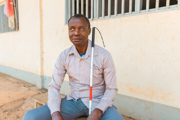 An African blind man with a cane sits on a bench