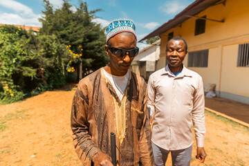 Two blind men standing in front of a house, one wearing a turban and sunglasses