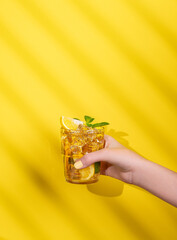 A hand holds a glass of iced tea with lemon and mint on a yellow background with shadow.