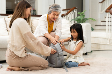 Happy Asian family preparing for school, helping daughter put books in school bag at home. Mother and grandmother take care of little cute girl to get ready for school at first day to school
