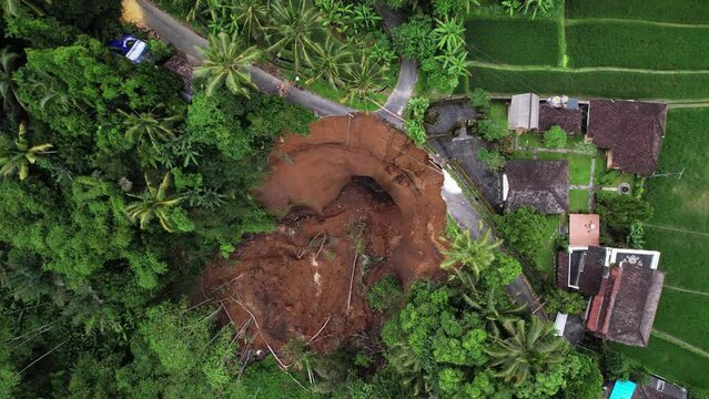 Top-down aerial shot of landslide aftermath, village road crossing destroyed, big mass of ground fall down. Unstable soil at steep slope of ravine was saturated by water from leaking subak channel