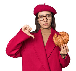 Chic Caucasian woman eating croissant in red coat and beret on studio background showing a dislike...