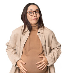 Pregnant young Caucasian woman showcasing maternity on studio background shrugs shoulders and open...