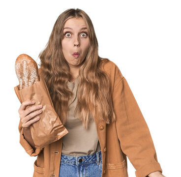 Blonde woman holding a loaf of bread in studio shrugs shoulders and open eyes confused.