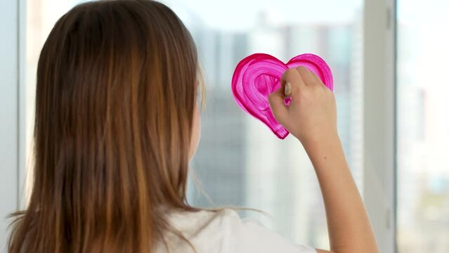Cute girl drawing heart on window with paintbrush, close-up