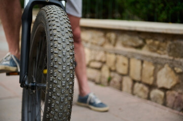 Naklejka premium Shallow tread of a bicycle tubeless tire. Selective focus on e-bike wheel and feet on pedals of a male cyclist in denim sneakers