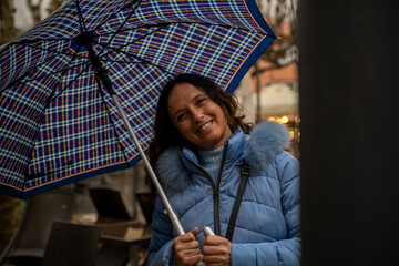 beautiful middle aged smiling woman walking in the rain with an umbrella in front of the tables of an cafe