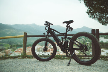 Fototapeta premium A full size shot of a modern black electric motor bike on the countryside road, parked by a wooden fence, against mountains background in the nature. Copy advertising space. Bike sharing city service.