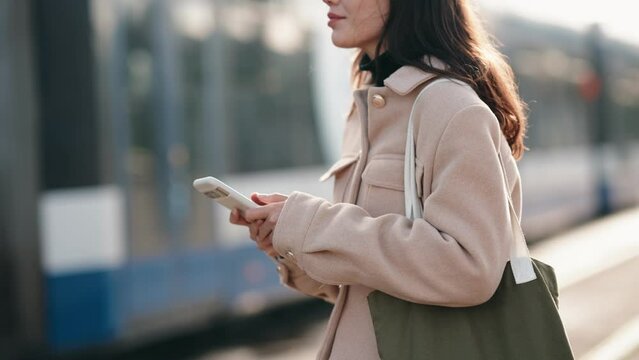 Capture of a faceless young woman browsing her smartphone at a tram station, depicting urban life and connectivity.