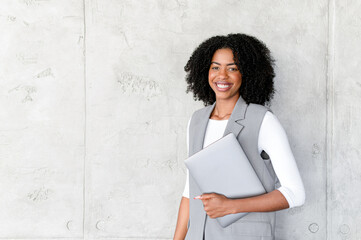 A poised African-American businesswoman stands confidently with her laptop, her smile exuding...