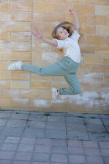 a girl wearing a white shirt and green pants is jumping in the air with a yellow brick wall behind her.