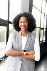 A poised African-American businesswoman with a cheerful demeanor stands arms crossed in a modern...