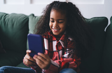 Smiling ethnic girl using smartphone on sofa in living room