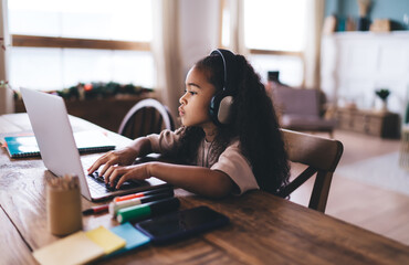 Adorable African American little girl sitting at wooden table typing on laptop and listening to music over headphones while looking at screen and doing home work in living room