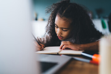 Serious African American curly haired female child studying and taking notes in notebook while sitting at wooden table with laptop against blurred background in living room - obrazy, fototapety, plakaty