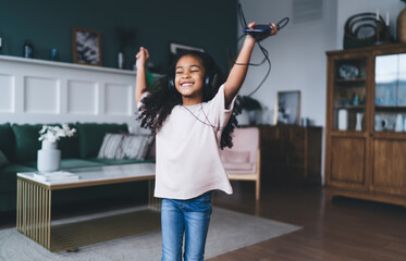 Carefree and cheerful African American girl in casual clothes playing music with smartphone while...