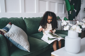 Concentrated African American girl, approximately 6-8 years old, sitting on a green sofa, intently...