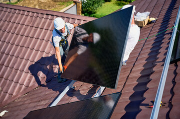 Workers building solar panel system on roof of house. Two men installers in helmets carrying photovoltaic solar module outdoors. Alternative, green and renewable energy generation concept.