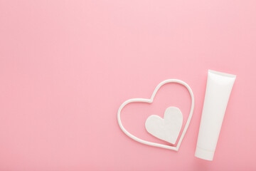 White heart shapes with cream tube on light pink table background. Pastel color. Care about female face, hands, legs and body skin. Closeup. Empty place for text. Top down view.