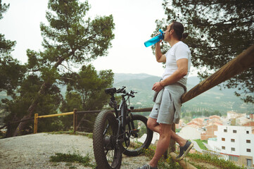 Caucasian young man cyclist in white t-shirt and sportswear, drinking water from sports bottle,...