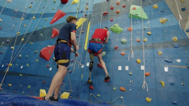 Low angle view of male climber with prothetic leg getting up wall in indoor gym while his belayer standing on floor and handling rope