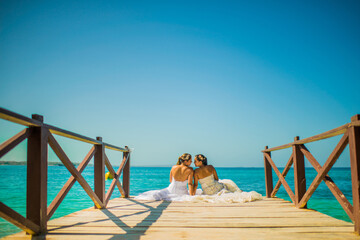 dos mujeres sentada mirandose frente a frente y usando vestidos de bodas en un muelle a la orilla...
