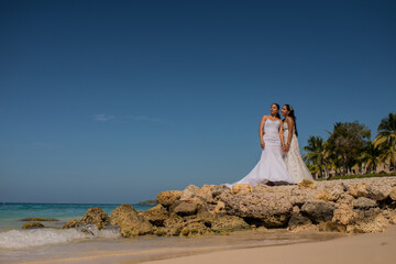 mujeres jovenes usando vestidos de boda y hablando a la orilla de la playa sobre unas rocas  - obrazy, fototapety, plakaty