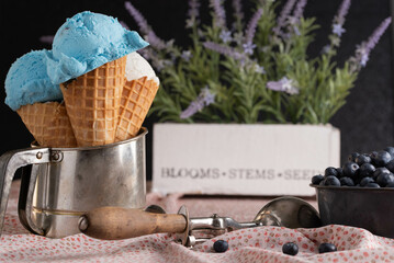 Blueberry and vanilla ice cream in waffle cone in an antique flour sifter on a floral-patterned tablecloth with fresh blueberries and a scoop.