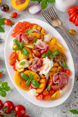 Caprese salad with mozzarella, tomatoes, prosciutto and grilled peaches on a white plate on a gray concrete background. Fresh summer salad. Top view.