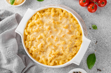 Mac and cheese in a white baking dish on a wooden board on a gray concrete background. American cuisine, comfort food. Copy space.