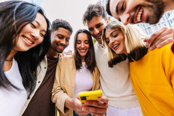 Group of young happy people using mobile phone at city street. Millennial diverse friends enjoying social media content on cellphone app. Technology lifestyle and youth community concept.