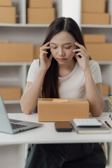 Freelance Asian woman wearing an apron uses laptop and boxes to take and check online orders to prepare packs to sell to customers. SME online business idea, vertical image