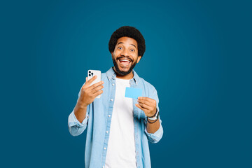 Full of excitement, young Brazilian man looks at the camera while holding a credit card and smartphone after a successful online transaction or receiving good financial news, isolated on blue