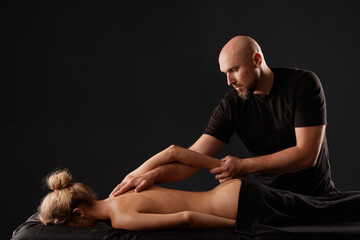 handsome male masseur doing a massage on a girl's hand in a black room, therapeutic relaxing massage concept - obrazy, fototapety, plakaty