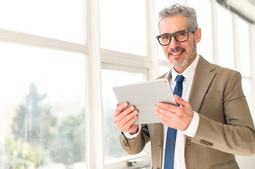 A senior businessman engages with modern technology, holding a tablet with an assured smile, showcasing how traditional business acumen blends with contemporary tools